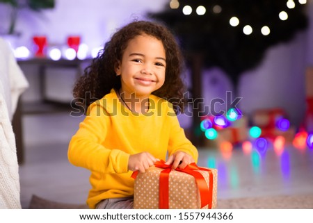 Happy little afro girl opening Christmas present in front of xmas tree at home, blurred background