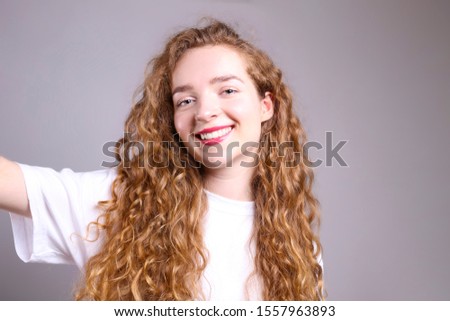 Young beautiful woman with long curly hair posing over isolated grey background. Portrait of teenage female model wearing white blank t-shirt, showing emotions. Close up, copy space.