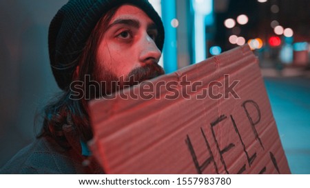 Side view of a bearded beggar with long hair asking for help with cardboard. Homeless concept.