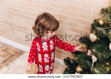 Beautiful little girl with curls in a red Christmas costume stands near the Christmas tree