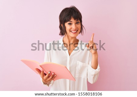 Young beautiful teacher woman reading book standing over isolated pink background surprised with an idea or question pointing finger with happy face, number one