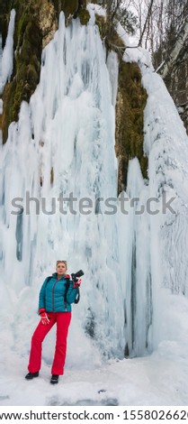 Portrait of woman traveler photographer in an blue jacket  and red  bologna pants with a camera in hand in the mountains on background of a frozen mountain lake. Photo travel concept. icy waterfall