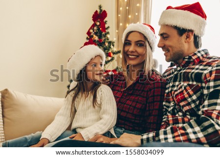 Christmas. Family. Happiness. Dad, mom and daughter in Santa hats are sitting on a couch at home near the Christmas tree, all are smiling