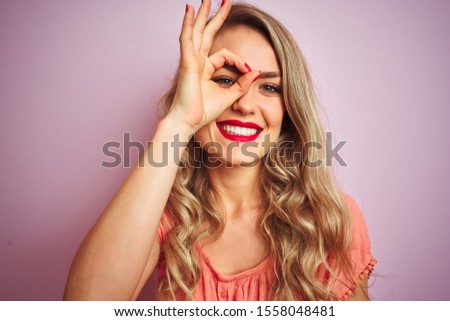 Young beautiful woman wearing t-shirt standing over pink isolated background with happy face smiling doing ok sign with hand on eye looking through fingers