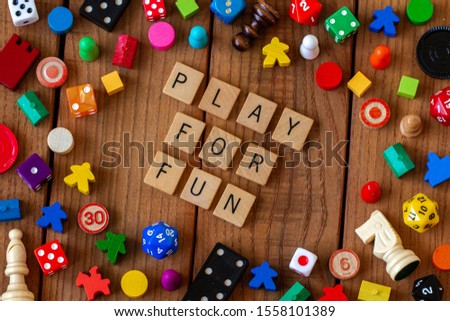 "Play For Fun" spelled out in wooden letter tiles. Surrounded by dice, cards, and other game pieces on a wooden background