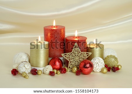Red and gold burning Christmas candles surrounded by natural potpourri elements and gold stars on a cream satin draped background