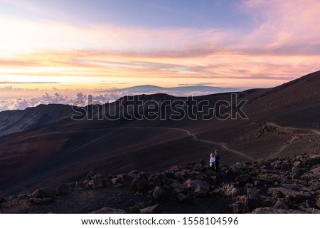 Couple watching a colorful sunrise on top of a big volcano with a view over the clouds. Shot on the Haleakala volcano summit on Maui, Hawaii USA.