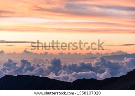Colorful sunrise on top of a big volcano with a view over the clouds. Shades of pink, purple and orange with the mountains' silhouette. Shot on the Haleakala volcano summit on Maui, Hawaii USA.