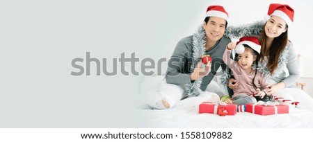 Asian family have father, mother and daughter with gray, pink casual wear and Santa hats. Sitting on a white bed with a red gift box And  small ball. Celebrate Christmas and Happy New Year. copy space