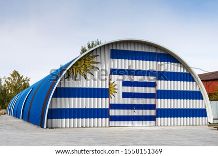 Close-up of the national flag of Uruguay
 painted on the metal wall of a large warehouse the closed territory against blue sky. The concept of storage of goods, entry to a closed area, logistics