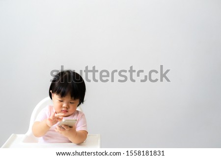 Adorable Asian baby girl is trying to use smartphone for video call camera or watching cartoon. Baby child girl is sitting on the highchair with white  background. Baby and Technology concept.