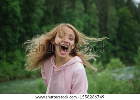 Funny portrait of amazing emotional young girl in pink casual clothes with long hair looking at the camera and grimacing on the hill with green forest background during spring in the mountains.