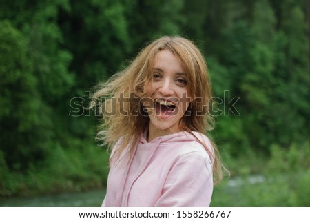 Funny portrait of amazing emotional young girl in pink casual clothes with long hair looking at the camera and grimacing on the hill with green forest background during spring in the mountains.