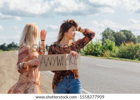 Two beautiful hippy girls hitchhiking and vote with a sign ANYWHERE on road. Copy space.