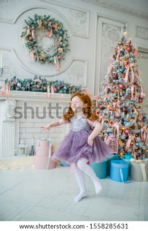 Cute funny little redhead child girl dance near decorated Christmas tree indoors. Merry Christmas and Happy Holidays.