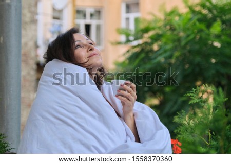 Beautiful mature woman relaxing on an open balcony with cup of coffee and dressed in white blanket, buildings and green trees background