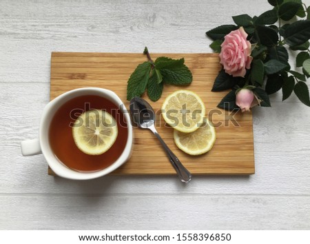 A still life on the light wooden background, a cup of tea with slices of lemon, spoon, mint leaves and a pale pink rose flower, flat lay, morning tea composition, top view