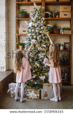 Two girls in a pink elegant dress is standing at the Christmas tree back, considering Christmas-tree decorations