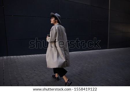 Fashionable young pretty curly woman with short haircut walking on city street in beige long trench, black trousers and wide grey hat, carrying takeaway coffee for her colleague