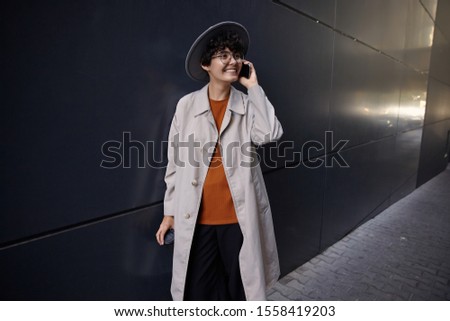Good looking smiling young curly brunette lady having pleasant conversation on phone while posing over black urban wall, wearing stylish clothes and wide grey hat