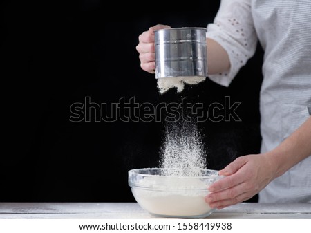 Chef sifts flour through a sieve in a glass bowl. Empty space for text. Isolated on dark background