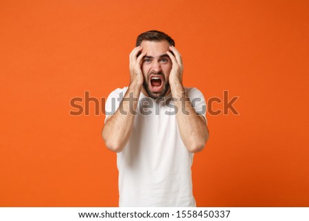 Shocked irritated young man in casual white t-shirt posing isolated on bright orange wall background, studio portrait. People lifestyle concept. Mock up copy space. Screaming, putting hands on head