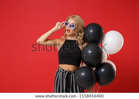Amazed young woman girl in black clothes, eyeglasses posing isolated on red background. International Women's Day birthday holiday party concept. Mock up copy space. Celebrating, holding air balloons