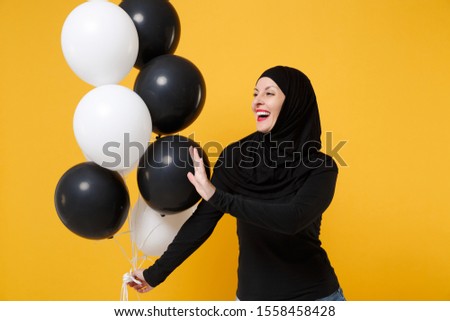 Arabian muslim woman in hijab celebrating holding black white air balloons isolated on yellow background studio portrait. Birthday holiday party people religious lifestyle concept. Mock up copy space