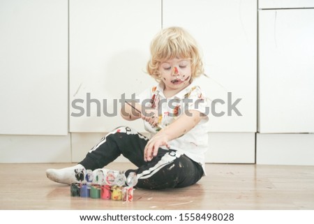 two year old boy with hands and face painted in colorful paints ready for more fun. dirty and happy kid sitting on the floor in white room background.