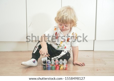 two year old boy with hands and face painted in colorful paints ready for more fun. dirty and happy kid sitting on the floor in white room background.