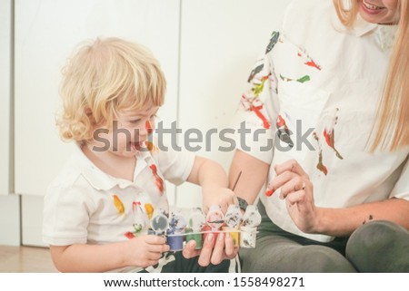 a cheerful mother and a joyful child sit on the floor in white t-shirts and paint each other with paints