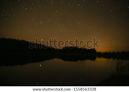 Dark night landscape by the lake. A black forest reflecting in the water and a sky full of stars. Orange hue and a haze