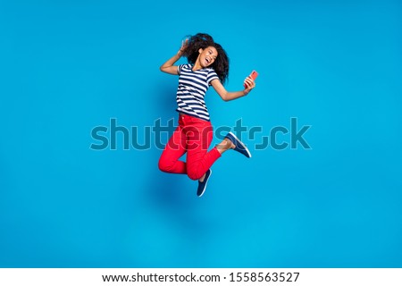 Full length body size turned photo of cheerful positive cute curly wavy pretty girlfriend jumping in striped t-shirt red trousers taking selfie isolated vivid blue color background