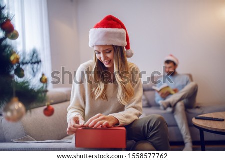 Smiling attractive caucasian woman in sweater and with santa hat on head sitting on sofa in living room next to christmas tree and packing gifts. In background her boyfriend sitting and reading book.