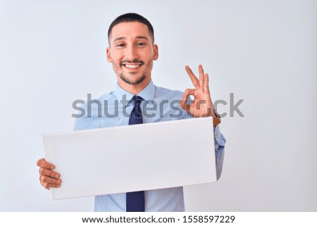 Young business man holding blank banner over isolated background doing ok sign with fingers, excellent symbol