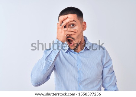 Young handsome business man standing over isolated background peeking in shock covering face and eyes with hand, looking through fingers with embarrassed expression.