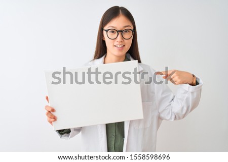 Young chinese dooctor woman wearing glasses holding banner over isolated white background with surprise face pointing finger to himself