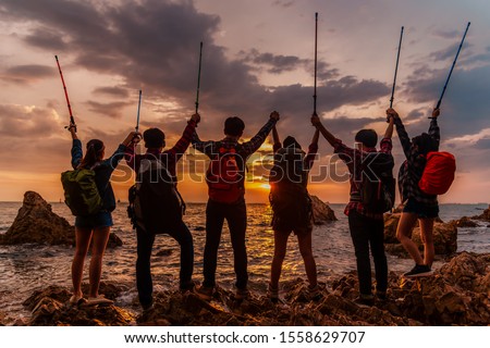 Group of asian hikers friends team with backpacks standing and hands up after success doing climbing activities at seaside. Travel, tourism, hiking and climbing concept.