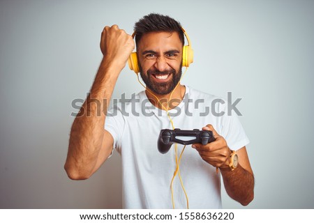 Arab indian gamer man playing video game using headphones over isolated white background annoyed and frustrated shouting with anger, crazy and yelling with raised hand, anger concept