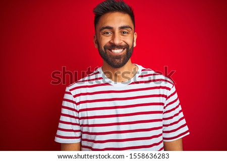 Young indian man wearing striped t-shirt standing over isolated red background with a happy and cool smile on face. Lucky person.