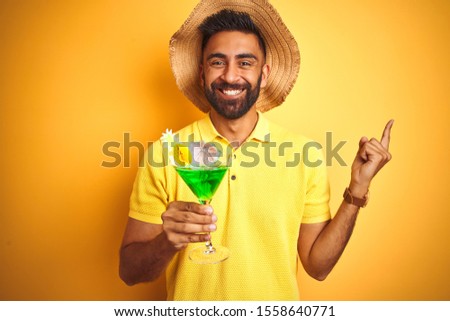 Indian man on vacation drinking cocktail wearing summer hat over isolated yellow background very happy pointing with hand and finger to the side