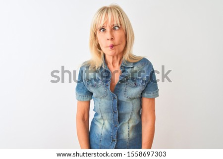 Middle age woman wearing casual denim shirt standing over isolated white background making fish face with lips, crazy and comical gesture. Funny expression.