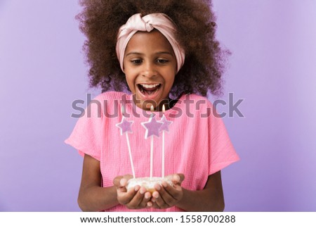 Image of a happy optimistic positive young african girl kid posing isolated over purple wall background holding birthday cake with candles.