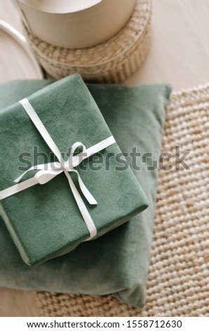Gift box wrapped in green packaging with a white ribbon, green pillow in a minimalistic festive interior