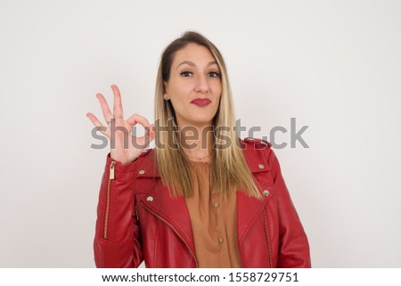 Glad attractive blonde woman shows ok sign with hand as expresses approval, has cheerful expression, being optimistic. Standing against white wall.