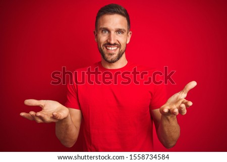 Young handsome man wearing casual t-shirt over red isolated background smiling cheerful offering hands giving assistance and acceptance.