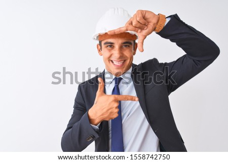 Young handsome architect man wearing suit and helmet over isolated white background smiling making frame with hands and fingers with happy face. Creativity and photography concept.