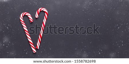 Two candy canes in shape of heart on black wooden background. Christmas, winter, new year concept. Snowfall drawing effect. Horizontal banner - Image