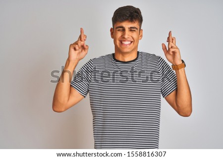 Young indian man wearing navy striped t-shirt standing over isolated white background gesturing finger crossed smiling with hope and eyes closed. Luck and superstitious concept.