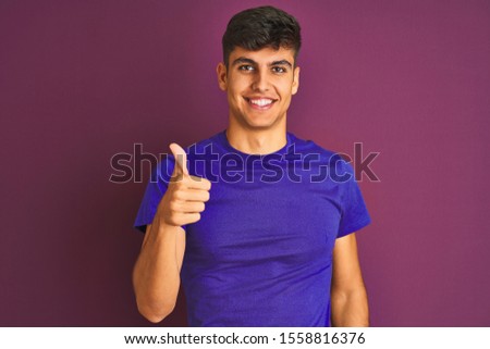 Young indian man wearing t-shirt standing over isolated purple background doing happy thumbs up gesture with hand. Approving expression looking at the camera showing success.